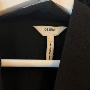 Svart kappa från Object - Elegant svart kappa från Object med långa ärmar och en stilren design. Perfekt för att ge en sofistikerad touch till din outfit. Kappan har en klassisk krage och en knappdetalj vid axeln.