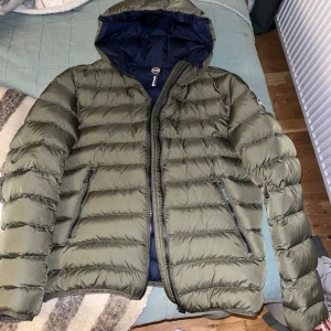 Olivgrön pufferjacka från Colmar - Säljer en olivgrön pufferjacka från Colmar med huva och dragkedja. Jackan är i toppskick utom en liten skavank vid baksidan på högerarmen, se sista bilen, men den är gömd så man ser den inte. Perfekt för kyligare väder.