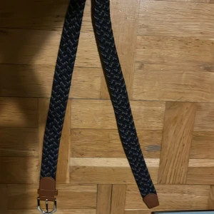 Svart och brunt flätat bälte - Snyggt flätat bälte i svart med bruna detaljer. Bältet har en stilren design med en silverfärgad spänne. Perfekt för att ge en extra touch till din outfit.