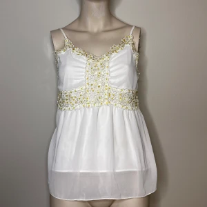 Vit linne från Vila - Säljer en söt vit linne från Vila med vackra spetsdetaljer och broderier i gult. Linnet har smala axelband och en luftig passform. Tillverkad i 100% polyester.