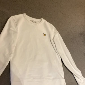 Vit tröja från Lyle & Scott - Säljer en stilren vit tröja från Lyle & Scott med deras ikoniska gula örn broderad på bröstet. Tröjan har långa ärmar och en klassisk rund halsringning. Perfekt för en avslappnad och snygg look.