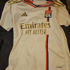 Vit fotbollströja från Adidas - Säljer en vit fotbollströja från Adidas med Olympique Lyonnais-logga och guldiga detaljer. Tröjan har korta ärmar och Aeroready-teknologi för optimal komfort. Den har även röd och blå rand längs sidan och sponsortryck på framsidan.