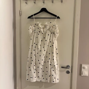 Vit prickig klänning med volangdetaljer - Charmig vit klänning med svarta prickar och volangdetaljer vid axlarna. Axelbandslös design som ger en luftig och somrig känsla. Perfekt för varma dagar och avslappnade tillfällen.