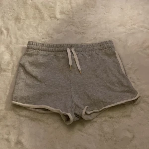 Gråa shorts från H&M - Säljer ett par grå shorts från H&M i ekologisk bomull. De har en elastisk midja med vit snörning och vita detaljer längs kanterna. Perfekta för en avslappnad stil.
