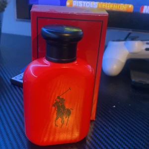 Röd parfymflaska från Ralph Lauren - Säljer en stilren röd parfymflaska från Ralph Lauren med den ikoniska polospelaren i svart på framsidan. Flaskan har en elegant svart kork och kommer i en matchande röd kartong. Perfekt för den som vill ha en klassisk och tidlös doft.