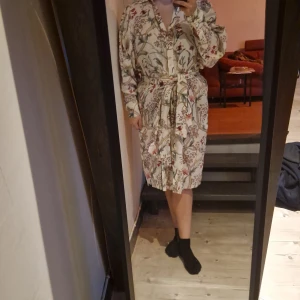 Blommig klänning med knytband - Charmig blommig klänning med knytband i midjan. Klänningen har långa ärmar och ett vackert mönster i olika färger. Perfekt för en avslappnad och stilfull look.