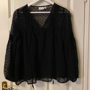 Svart blus från VILA - Säljer en elegant svart blus från VILA med prickigt mönster. Blusen har en v-ringad design och långa ärmar i ett transparent material. Perfekt för en stilfull look.