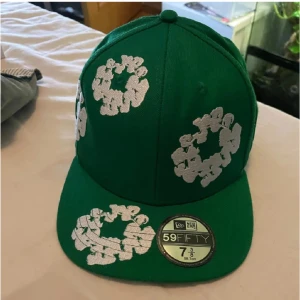 Grön keps från Denim tears - Snygg grön keps från Denim tears med broderade vita kransar. Modellen är 59/50 och har en klassisk passform. Perfekt för att ge din stil en unik touch.