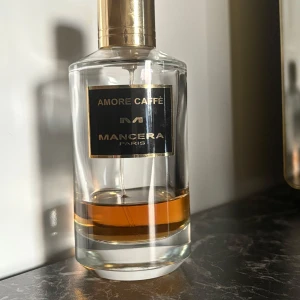 Amore Caffè parfym från Mancera - Elegant parfymflaska från Mancera med doften Amore Caffè. Flaskan är genomskinlig med en gyllene kork och en svart etikett med guldtext. Perfekt för den som älskar en djup och rik doftupplevelse. Ursprungligen 120ml