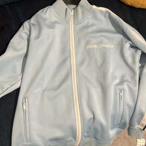 Ljusblå jacka från Palm Angels - Säljer en stilren ljusblå jacka från Palm Angels med dragkedja och vita detaljer. Jackan har långa ärmar med vita ränder längs sidorna och två fickor framtill. Perfekt för en avslappnad look.