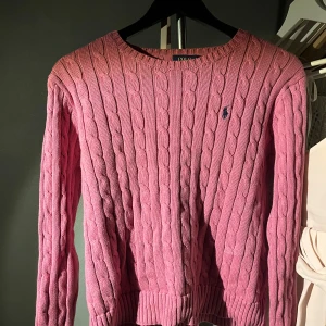 Rosa stickad tröja från Ralph Lauren - Säljer en snygg rosa stickad tröja från Ralph Lauren med kabelstickat mönster och den klassiska loggan på bröstet. Tröjan har långa ärmar och en rund halsringning. Perfekt för en stilren look!