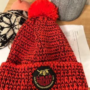 Röd och svart mössa från Luleå Hockey - Snygg röd och svart stickad mössa från Luleå Hockey med en stor tofs på toppen. Mössan har ett broderat emblem framtill med klubbens logga och årtal. Perfekt för att hålla värmen under kalla dagar.