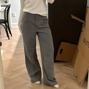 Grå kostymbyxor från Claire Rose x NA-KD - Snygga grå kostymbyxor från Claire Roses kollektion med NA-KD med hög midja och rak passform. Perfekta för en stilren look. Byxorna har hällor för bälte och en diskret dragkedja framtill. Det finns även gummiband inne i midjan så att det går att justera storleken. Storlek 34