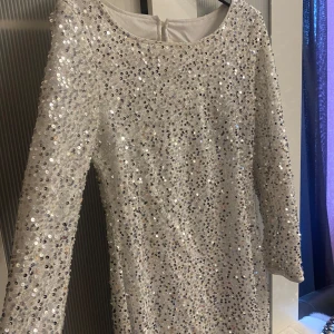 Vit paljettklänning - Säljer en glittrig vit klänning med paljetter. Klänningen har långa ärmar och en elegant passform som passar perfekt för festliga tillfällen. Den är i en klassisk stil med en rund halsringning.
