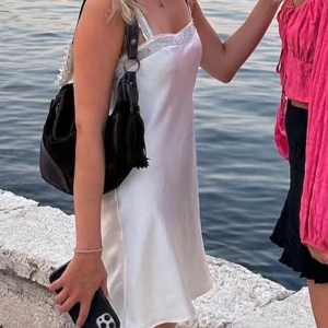 Vit axelbandslös klänning med spetsdetaljer - Elegant vit satin klänning med glitter/strass detalj längs urringningen. Perfekt för en stilren look, oavsett om du bär den ensam eller med en jacka. Klänningen har en mjuk och glansig yta som ger en lyxig känsla.    Sommar, fest, Stockholmsstil, midsommar, glitter, strass, paljetter 