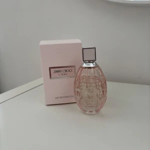 Jimmy Choo L'Eau Eau de Toilette - Elegant och feminin parfym från Jimmy Choo i en vacker ljusrosa flaska med ett mönstrat glas. Flaskan har en silverfärgad hals och en fyrkantig kork. Perfekt för den som älskar en fräsch och blommig doft.