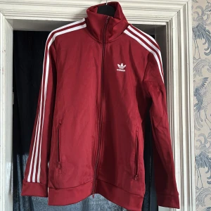 Röd träningsjacka från Adidas - Säljer en snygg röd träningsjacka från Adidas med de klassiska tre vita ränderna längs ärmarna. Jackan har dragkedja framtill och två fickor. Perfekt för en sportig look eller avslappnad stil.