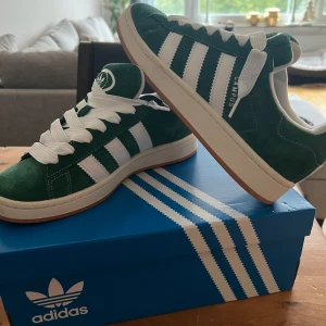Adidas campus 00 - Snygga gröna Adidas Campus sneakers i mocka med vita detaljer och snörning. De har den klassiska tre-randiga designen på sidorna och en vit sula. Perfekta för en stilren och sportig look.