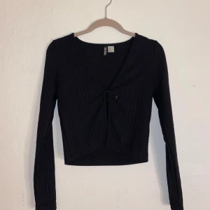 Svart ribbad kofta från H&M - Snygg svart ribbad topp från H&M med långärmad design och knappar framtill. Perfekt för en stilren look. Passar bra till både jeans och kjol.