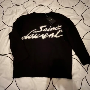 Svart tröja från Saint Laurent - Säljer en stilren svart tröja från Saint Laurent med vit text på framsidan. Tröjan har långa ärmar och en klassisk passform. Perfekt för en trendig look.