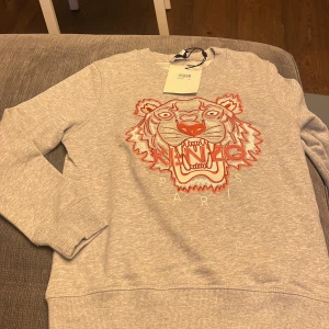 Grå oanvänd sweatshirt från Kenzo Kids - Säljer en grå sweatshirt från Kenzo Kids med ett coolt tigertryck i rött och vitt på framsidan. Tröjan har långa ärmar och en rund halsringning. Perfekt för en stilren och bekväm look.