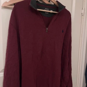 Vinröd tröja med dragkedja från Polo Ralph Lauren - Säljer en stilren vinröd tröja från Polo Ralph Lauren med halv dragkedja och broderad logga på bröstet. Tröjan har en klassisk design med långa ärmar och ribbade muddar. Perfekt för en avslappnad men ändå snygg look.