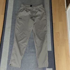 Beige träningsbyxor från Under Armour - Säljer ett par beige träningsbyxor från Under Armour. Byxorna har en elastisk midja med snörning och är perfekta för träning. De har en avslappnad passform och är tillverkade i ett lätt syntetmaterial. Har använt de 1 gång!Vill få bort den så fort som möjligt! Snabbast får!!