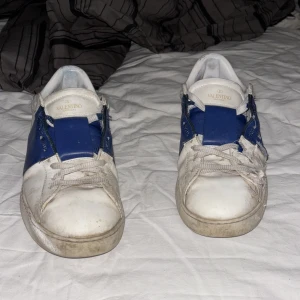 Valentino sneakers i vitt och blått - Säljer ett par Valentino sneakers i vitt med blå detaljer. Skorna har snörning och en klassisk design med rund tå. Perfekta för en stilren look. Defekt på vänster skon. Kom med bud!! INTE RIKTIGA PRISET!!!