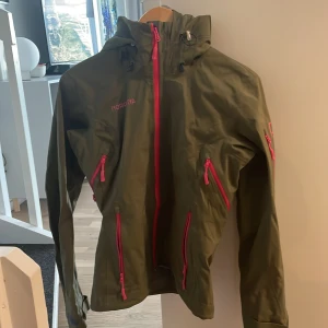 Grön vindjacka från Norrøna - Säljer en grön vindjacka från Norrøna med rosa dragkedjor och justerbar huva. Jackan har flera praktiska fickor med dragkedja och kardborrejustering vid ärmsluten. Perfekt för utomhusaktiviteter och äventyr.