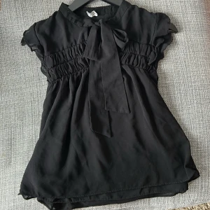 Svart blus med knytband - Säljer en elegant svart blus med knytband vid halsen. Blusen har korta ärmar och smockdetaljer vid bysten som ger en fin passform. Perfekt för en stilren look. Från Zaras tråd collection. 