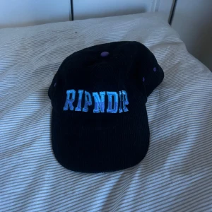 Svart manchesterkeps från Ripndip - Snygg svart keps i manchester från Ripndip med broderad text i blått och lila framtill. På sidan finns en broderad katt. Perfekt för en cool och avslappnad stil.