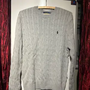 Säljer en stilren grå stickad tröja från Polo Ralph Lauren med kabelstickat mönster. Tröjan har långa ärmar och en broderad logotyp på bröstet. Perfekt för kyligare dagar. OBS! Priset är inte hugget i sten. Hör av er vid minsta fundering 🍾