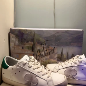 The gallerist skor storlek 38 - Snygga vita sneakers från The Gallerist med gröna detaljer på hälen och ett unikt klövermotiv på sidan. Skorna har snörning och en svart sula med bra grepp. Perfekta för en stilren look. Nästan helt nya, använda ca 4 gånger ute men dem är tvättade!