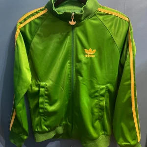 Grön sporttröja från Adidas - Säljer en grön träningsjacka från Adidas(inte helt hundra om den är äkta) med guldiga detaljer och dragkedja. Jackan har långa ärmar och ribbade muddar. Storlek M(ganska liten) , pris kan diskuteras!