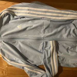 Ljusblå träningsjacka från Adidas - Säljer en ljusblå träningsjacka från Adidas med klassiska vita ränder längs ärmarna. Jackan har dragkedja framtill och Adidas-logga på bröstet. Perfekt för sportiga aktiviteter eller en avslappnad stil.