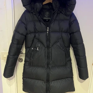 Svart dunjacka från Cedrico - Säljer en svart dunjacka från Cedrico med huva och dragkedja. Jackan har en stilren design med praktiska fickor framtill. Perfekt för kalla dagar när du vill hålla dig varm och bekväm.