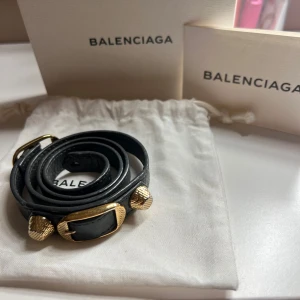 Svart armband från Balenciaga - Elegant svart armband från Balenciaga i skinn med guldfärgade detaljer. Armbandet har en justerbar rem och är dekorerat med tre guldfärgade nitar. Perfekt för att ge en lyxig touch till din stil. Nyskick med inga slitage!❤️