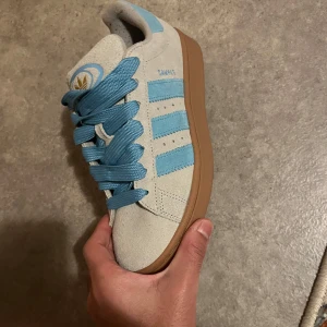 Adidas Campus sneakers i ljusblått och beige - Snygga Adidas Campus sneakers i en fräsch kombination av ljusblått och beige. Skorna har en klassisk design med tre ränder på sidan och en gummisula i brunt. Perfekta för en avslappnad stil.