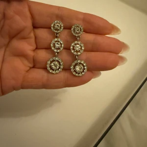 Glittriga örhängen med stenar - Snygga örhängen i silverfärg med tre rader av glittrande stenar. Perfekta för att ge en extra touch av glamour till din outfit. De har en elegant design med runda stenar i olika storlekar.