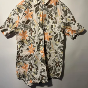 Kortärmad skjorta lager 157 - Vit skjorta med ett blommigt mönster i orange, brunt och grönt. Skjortan har korta ärmar och knappar framtill. Mycket bra skick, storlek M.