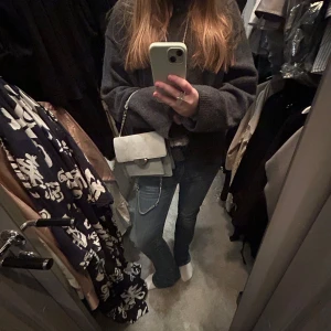 Grå mocka axelväska med kedja - Snygg grå axelväska i mocka med en stilren design. Väskan har en silverfärgad kedja som axelrem och ett lock med en metallspänne framtill. Perfekt för att ge en elegant touch till din outfit. Har en liten fläck som syns på bilden men inget man tänker på!