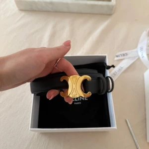 Svart bälte från Celine - Snyggt svart bälte från Celine med ett elegant guldfärgat spänne i form av deras ikoniska logotyp. Perfekt för att ge en lyxig touch till din outfit. Kommer i originalförpackning med dustbag och certifikat. Använt cirka 2 gånger, finns en liten repa men är inget som syns om man inte tittar jättenogrant och nära.  Modell mellan storlek 75. Ordinarie pris 6300 