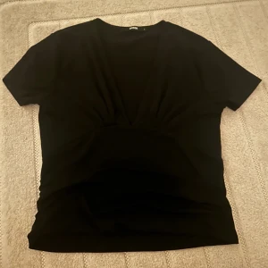 Svart v-ringad topp - Säljer en stilren svart topp med v-ringning och korta ärmar. Toppen har en åtsittande passform med rynkade detaljer vid midjan som ger en smickrande silhuett. Perfekt för en elegant look. (Andra bilden är inte min)