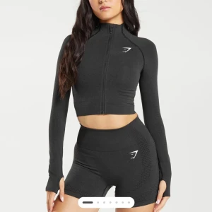 Svart träningsjacka från Gymshark - Säljer en svart ny croppad träningströja från Gymshark med dragkedja framtill. Jackan är i kortare modell och har långa ärmar med tumhål. Perfekt för träning med sin tighta passform och sömlösa design. Glömde göra retur, tags kvar på plagget!