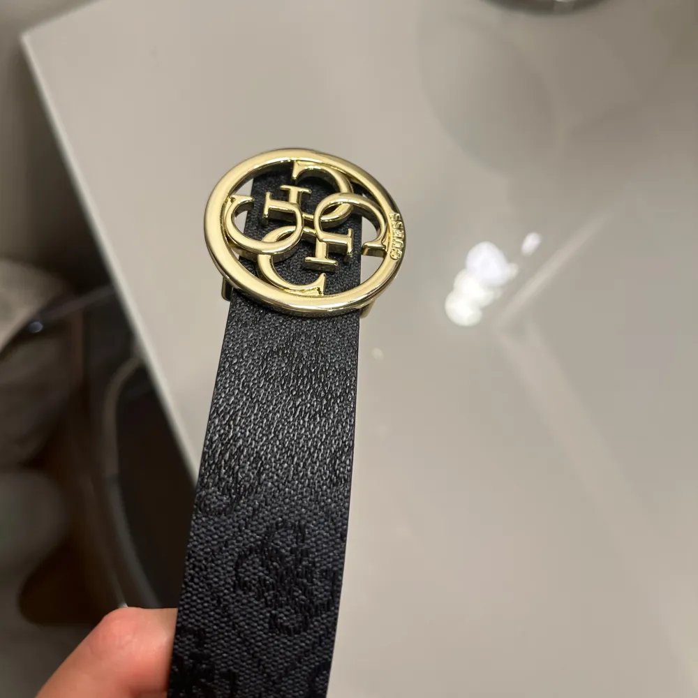 Snyggt vändbart bälte från Guess med ett elegant mönster. Ena sidan är brun med logotypmönster och den andra är svart. Bältet har ett stort, runt guldfärgat spänne med Guess-logotypen. Perfekt för att ge din outfit en lyxig touch. Oanvänd . Asusteet.