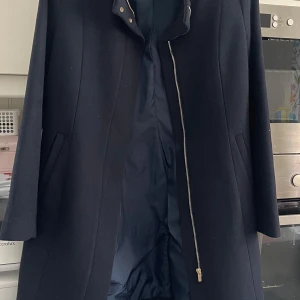 Svart kappa med dragkedja - Elegant svart kappa med stilren design och dragkedja framtill. Kappan har en hög krage och dolda fickor på sidorna. Perfekt för kyligare dagar. Använd endast en gång vid speciellt tillfällen 