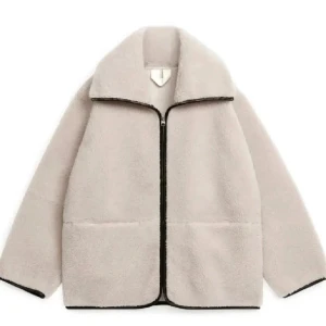 Teddyjacka  - Mysig beige teddyjacka med svart dragkedja och detaljer. Jackan har en hög krage och långa ärmar, perfekt för kyligare dagar. Den har en avslappnad passform och stora fickor framtill.