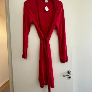 Röd långärmad klänning med knytband - Snygg röd långärmad klänning med knytband i midjan. Perfekt för att skapa en elegant och avslappnad look. Klänningen har en öppen design framtill och är tillverkad i ett mjukt material.