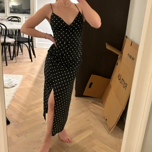 Svart prickig klänning med slits - Snygg svart klänning som går omlott med vita prickar och smala axelband. Den har en v-ringad design och en slits på sidan. Baktill finns en elegant snörning som ger en fin detalj. Endast använd en gång, som ny!