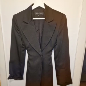 Svart trenchcoat från Shelli Segal - Elegant svart trenchcoat från Shelli Segal med klassisk design och knytband i midjan. Köpt i New York. Garderobsrensning så kika gärna på övriga auktioner. 